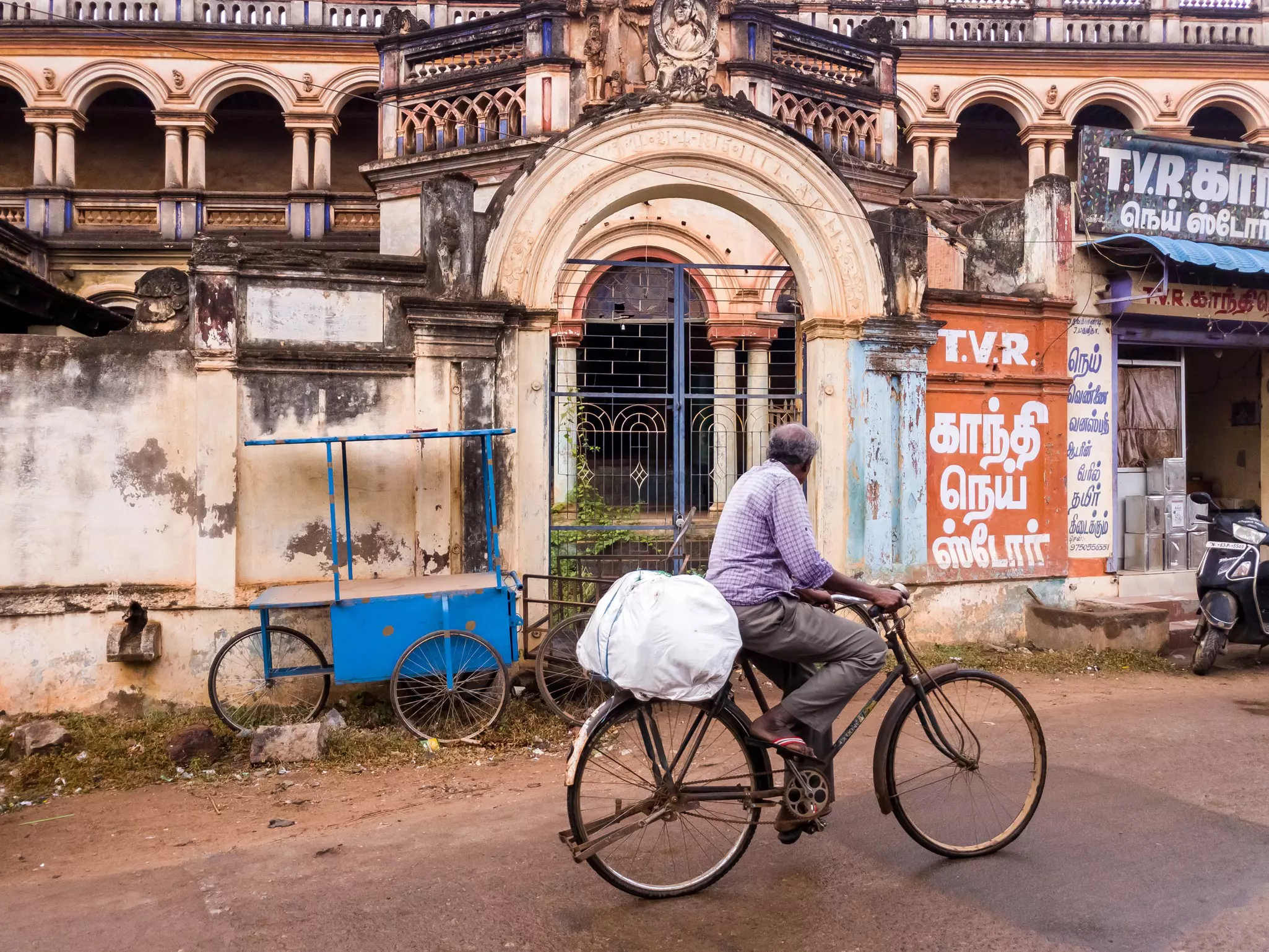 A man on a bicycle is seen from behind pedaling past a dilapidated mansion behind a gate.