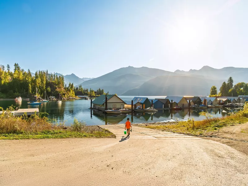 A young boy with a fishing rod and tackle box walks on a dirt road toward a bay boat ramp and boathouses along a lake on a sunny day.