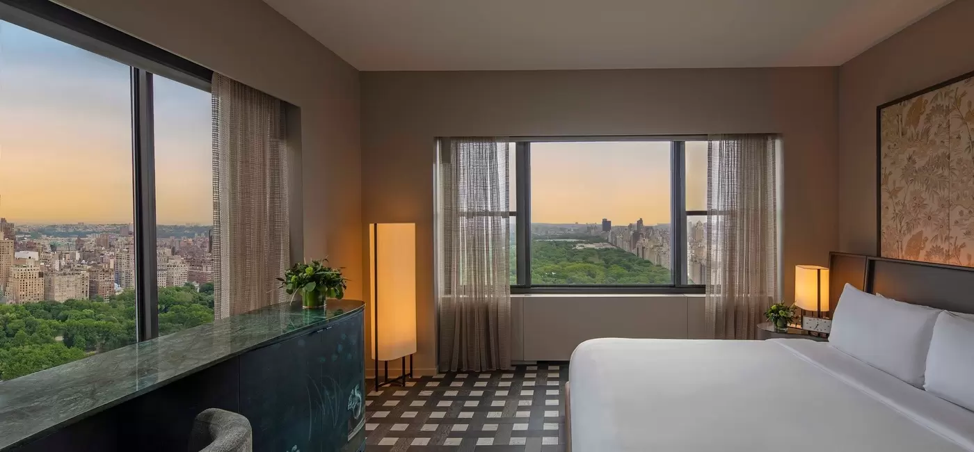 A white bed in a large corner hotel room; through the windows, the background is of trees and a city skyline.