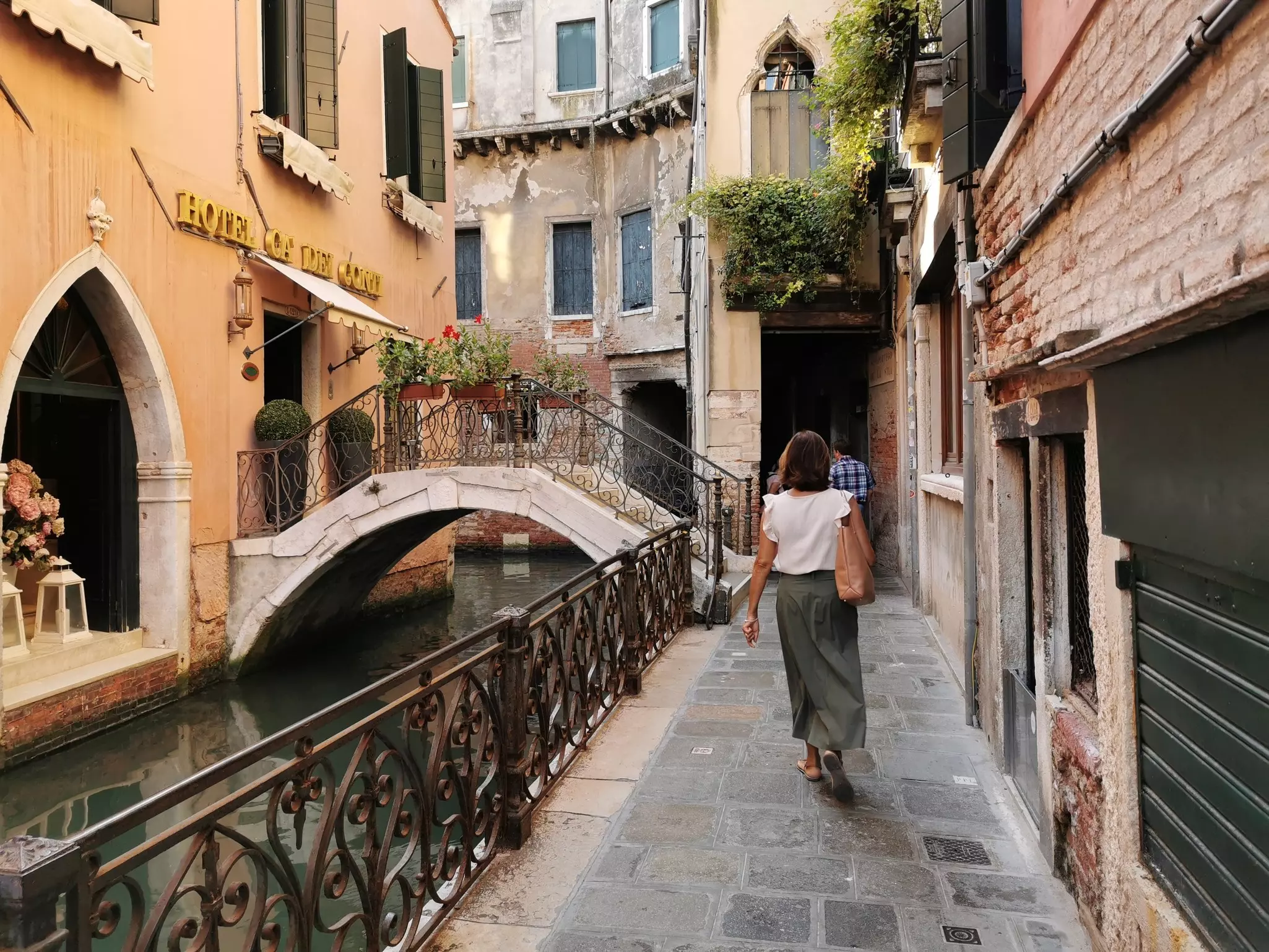 A woman walks along a narrow cobblestone alley beside a canal, passing historic Venetian buildings with aged facades and shuttered windows