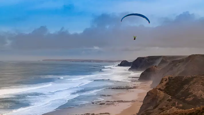 A paraglider flying off cliffs above an ocean beach.