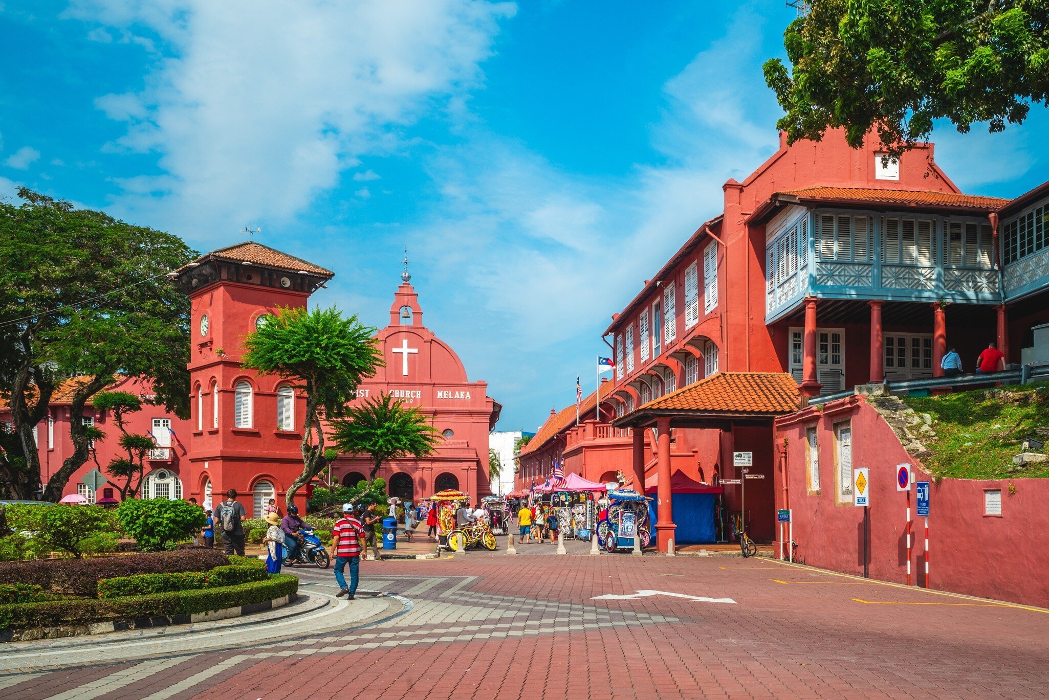 Red colonial Stadthuys on Melaka's Dutch Square beside a clock tower and Christ Church, with trishaws and pedestrians