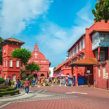 Red colonial Stadthuys on Melaka's Dutch Square beside a clock tower and Christ Church, with trishaws and pedestrians