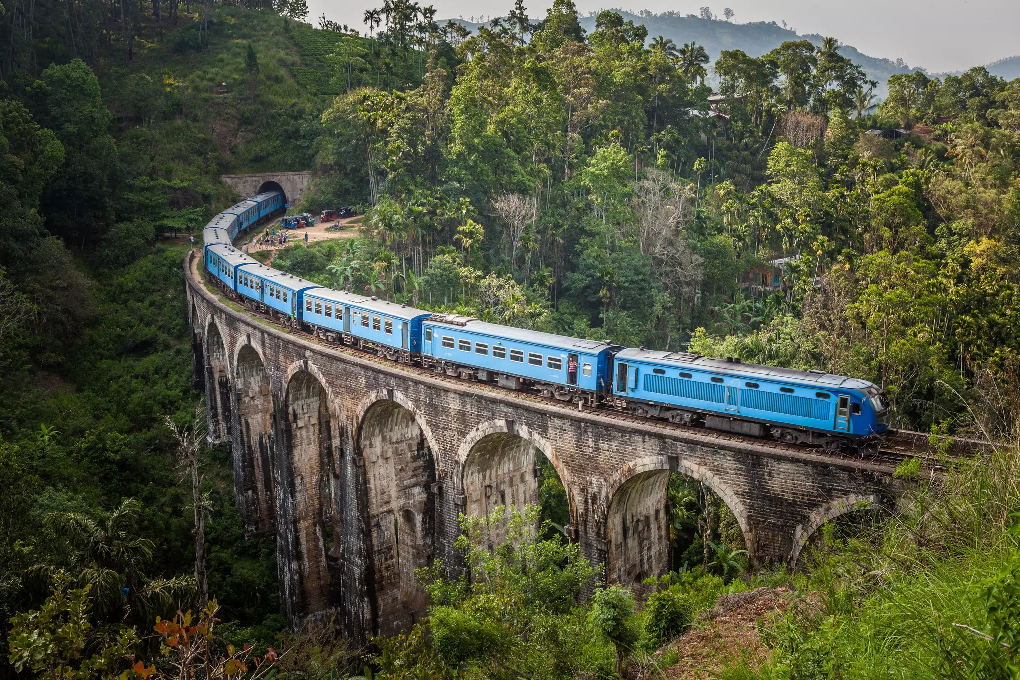 A blue train passes over an arched bridge surrounded by jungle near Ella in Sri Lanka.