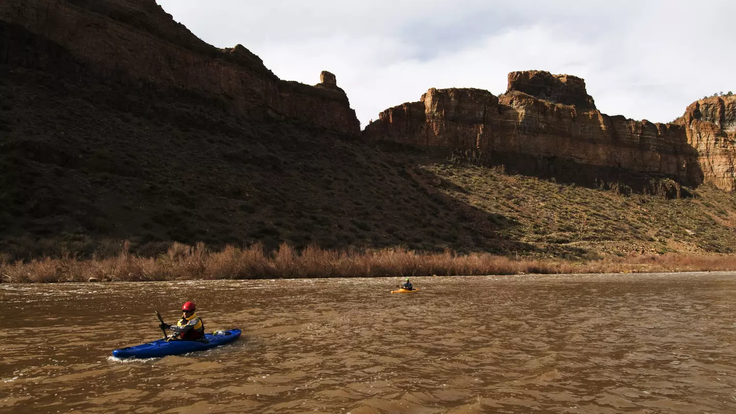Kayakers paddle their boats on the Salt River against rocky mountains in shadow