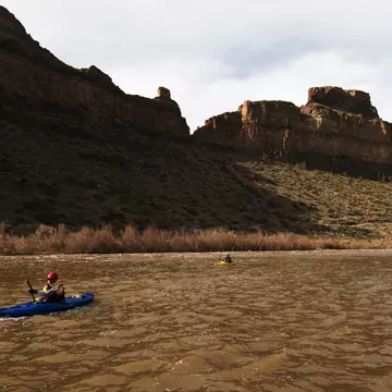 Kayakers paddle their boats on the Salt River against rocky mountains in shadow