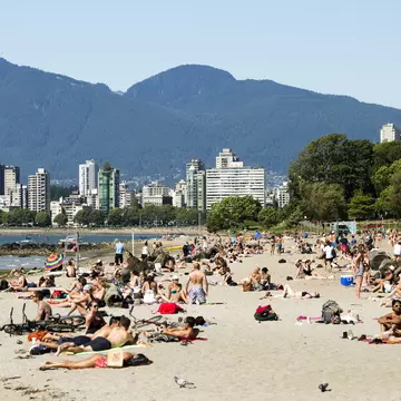 People sunbathe and chat in groups on a beach near an urban area.