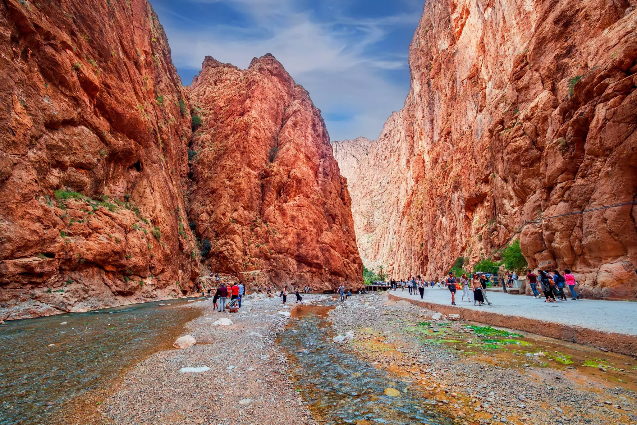 Hikers follow a trail through a river gorge with steep red-rock cliffs.