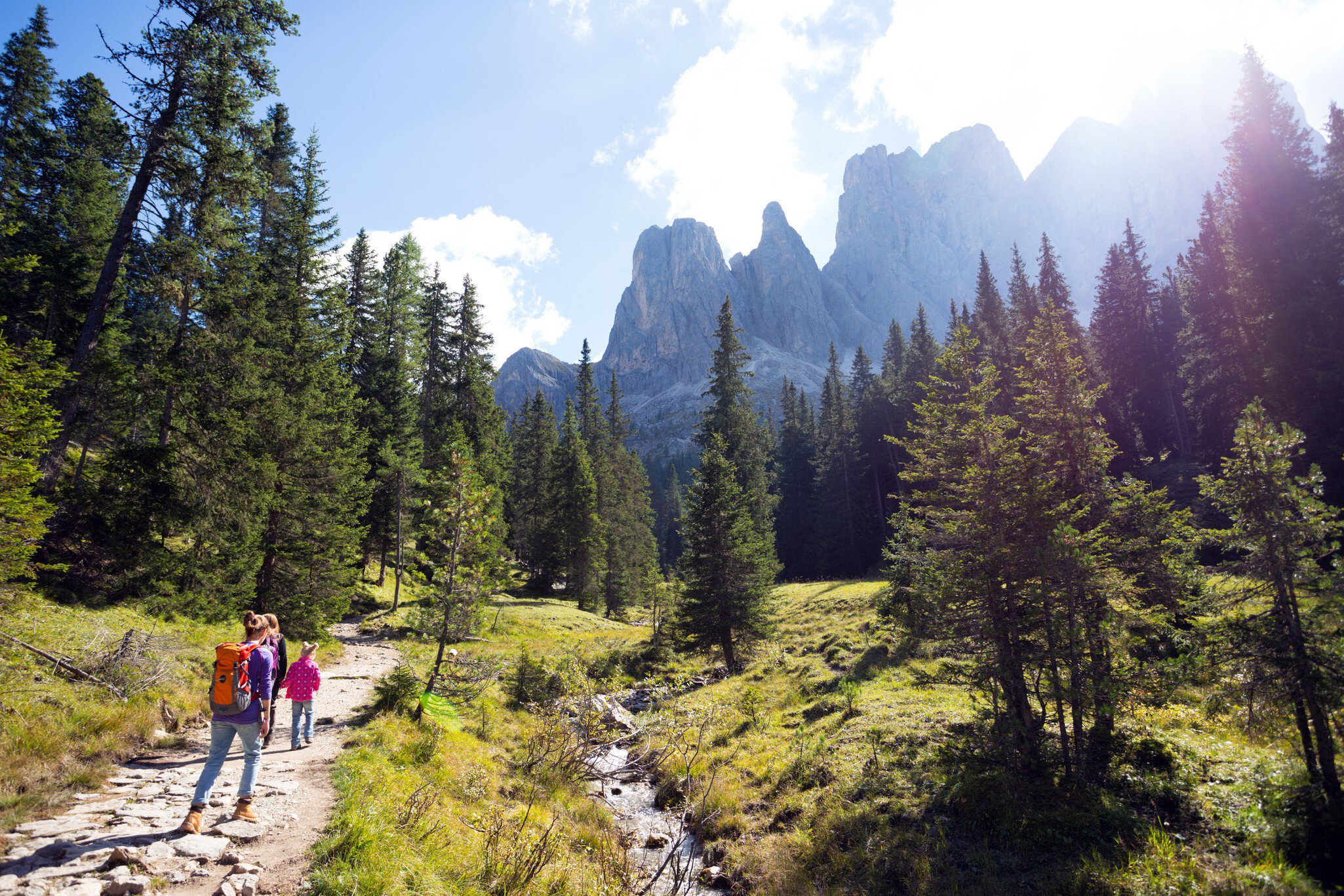 Two adults and a child hike along a path beside a stream in a mountainous region of Italy, with tall trees and mountains by the path.