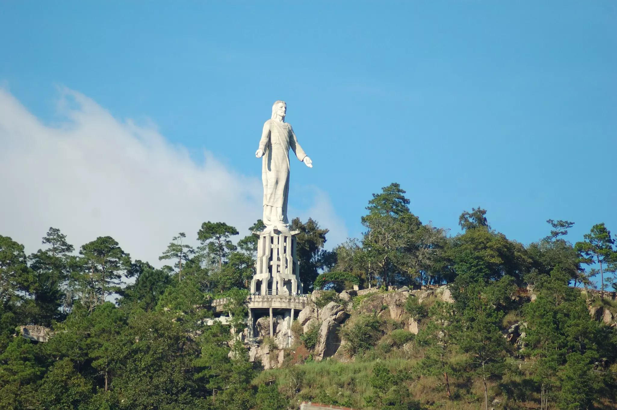 A stone statue of a man with his arms held open rises over the forests on a hilltop.
