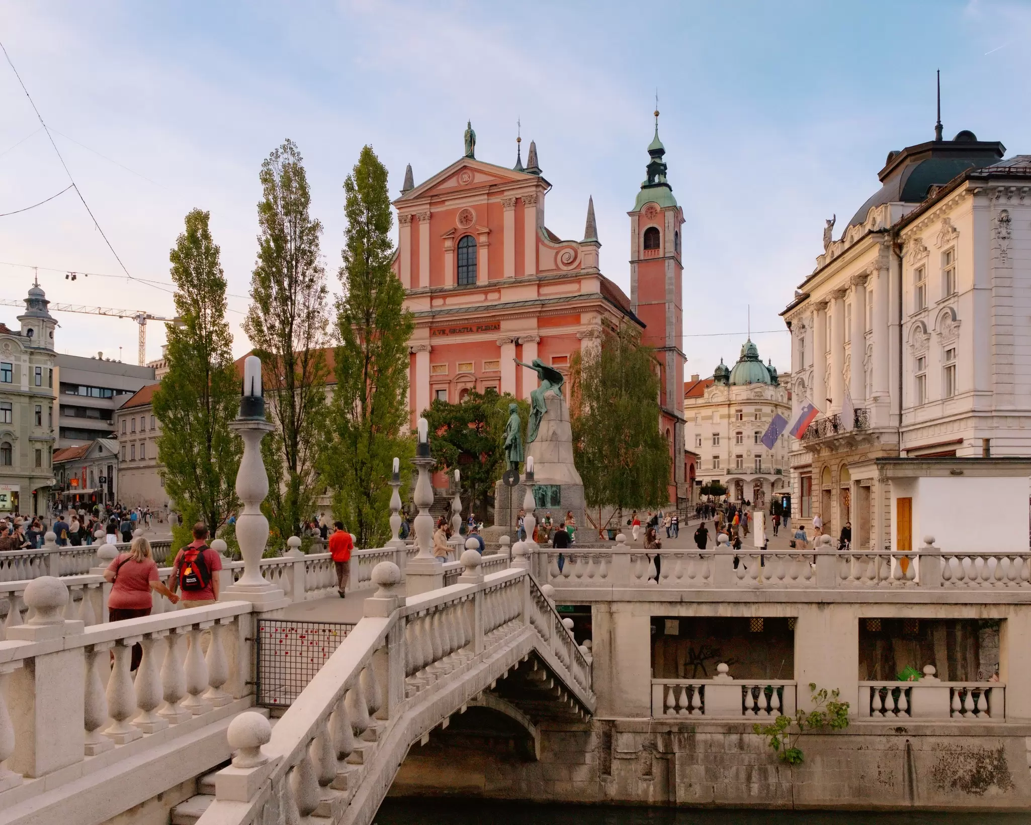 People walking along an ornate stone bridge in a city with large buildings in the background.