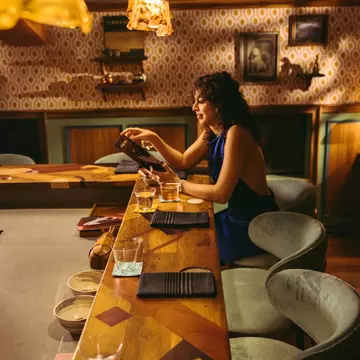 A lone woman sits a chef counter in a dimly lit restaurant holding a menu.