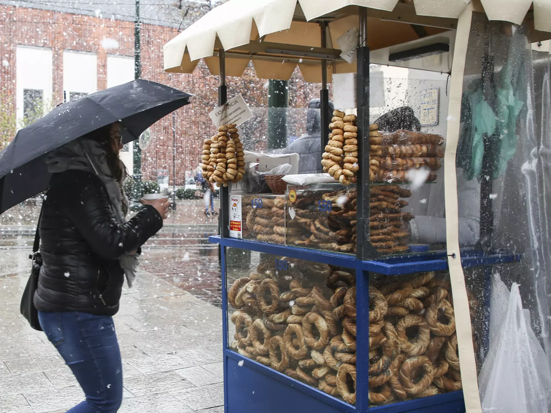 A woman holding a cup under an umbrella during a snowstorm waits at a blue obwarzanek stall in Kraków.