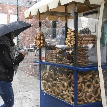 A woman holding a cup under an umbrella during a snowstorm waits at a blue obwarzanek stall in Kraków.
