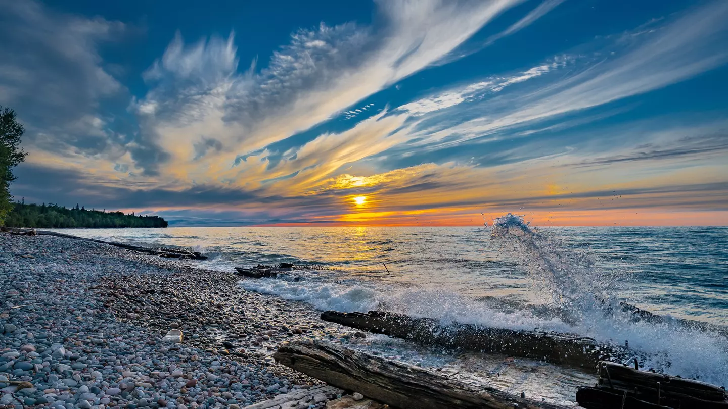 The sun sets over the lake; the shore is littered with driftwood