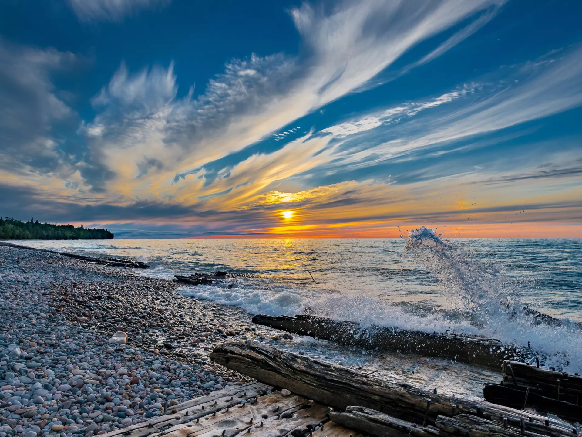 The sun sets over the lake; the shore is littered with driftwood