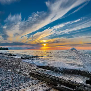 The sun sets over the lake; the shore is littered with driftwood