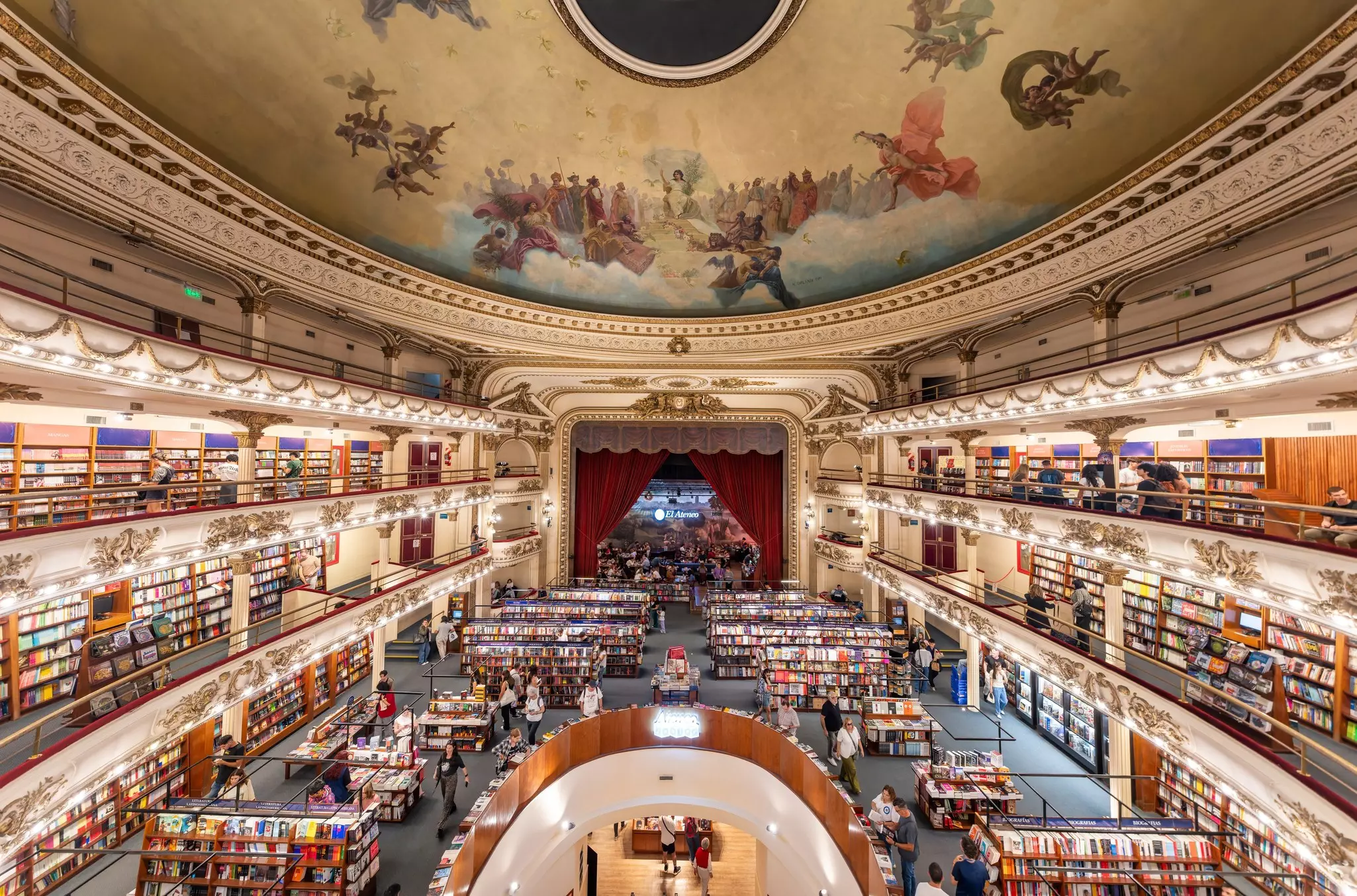 A vast oval room of a former theater lined with bookshelves. A stage, with red curtains, is now home to a cafe.
