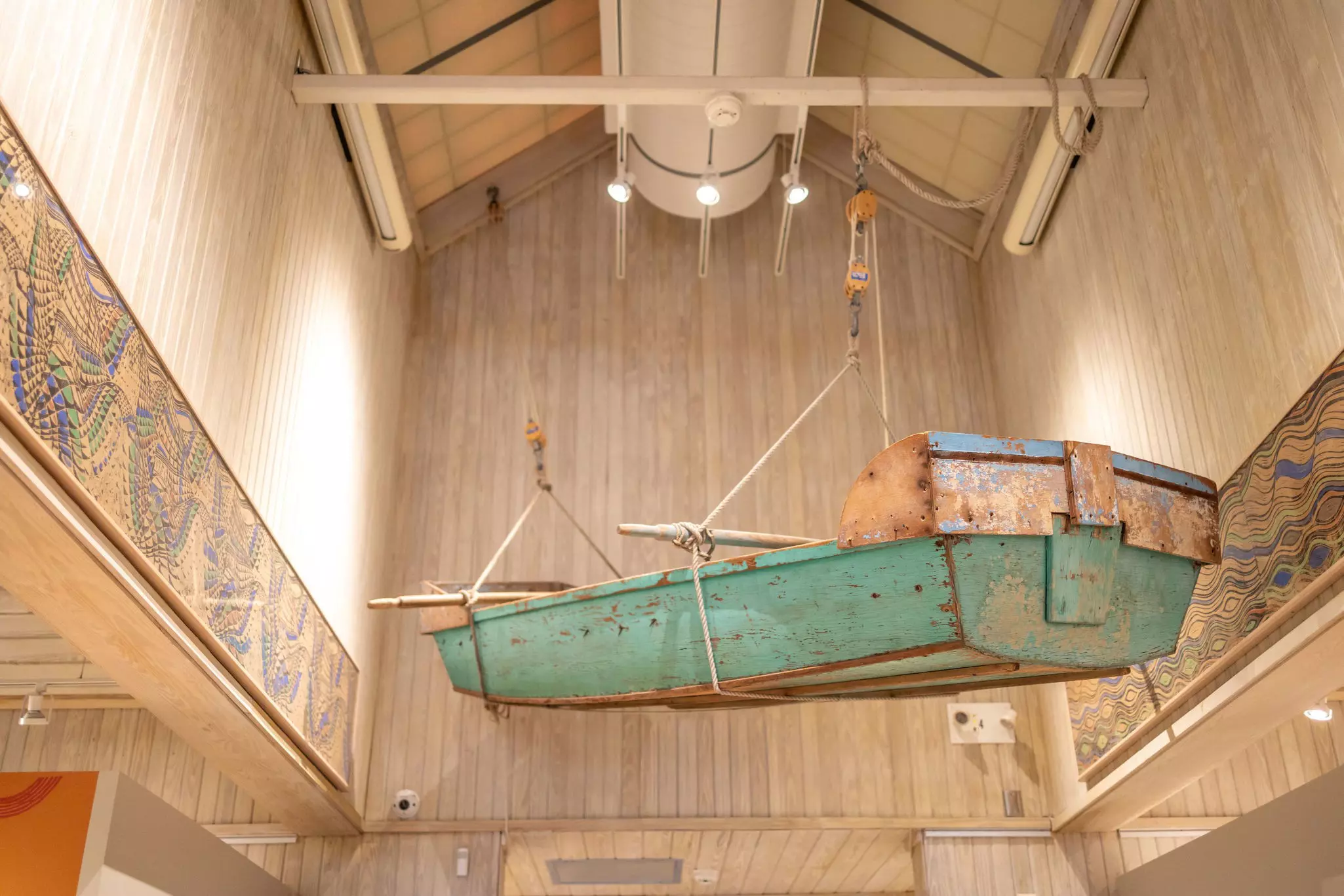 A rowboat hangs in the atrium of a museum gallery. Paintings are displayed on the gallery’s walls.