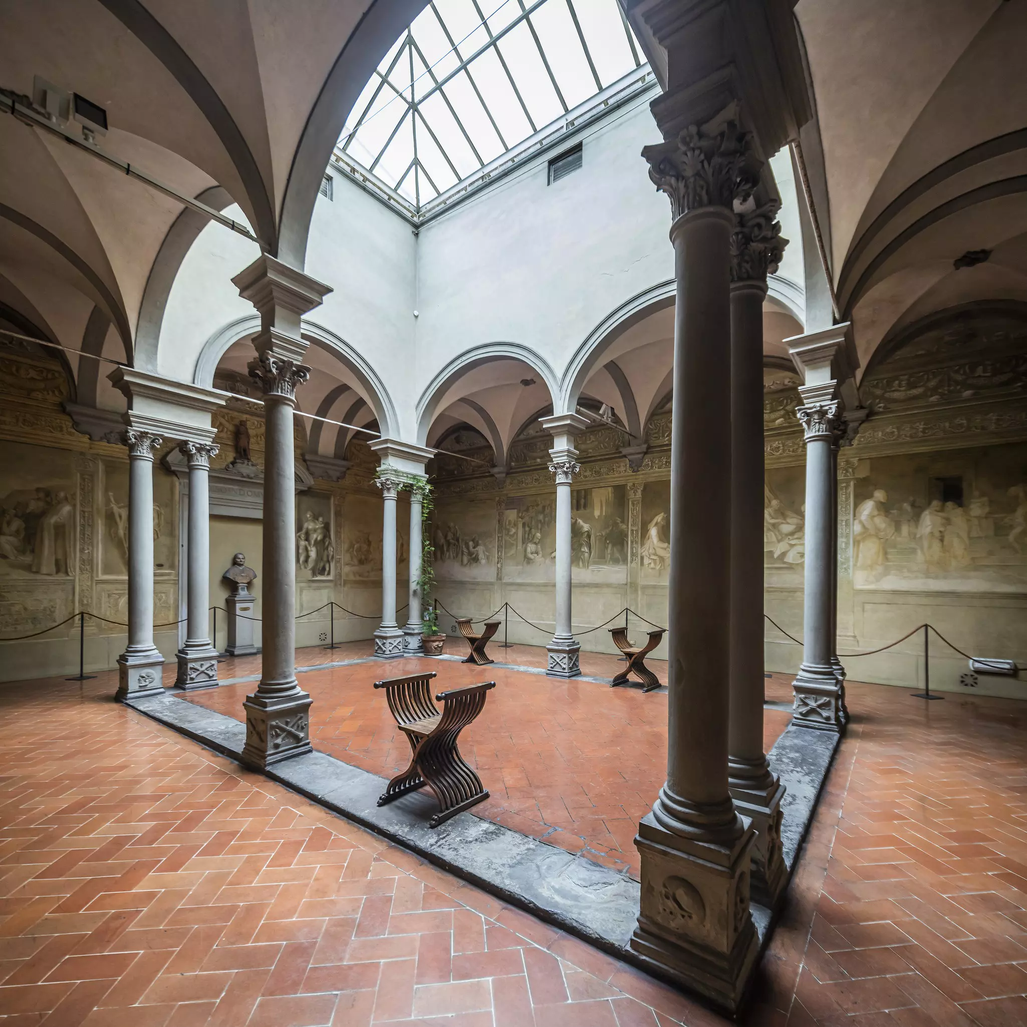 A cloister with a glass roof and a red tiled floor lined with black-and-white drawings of people.