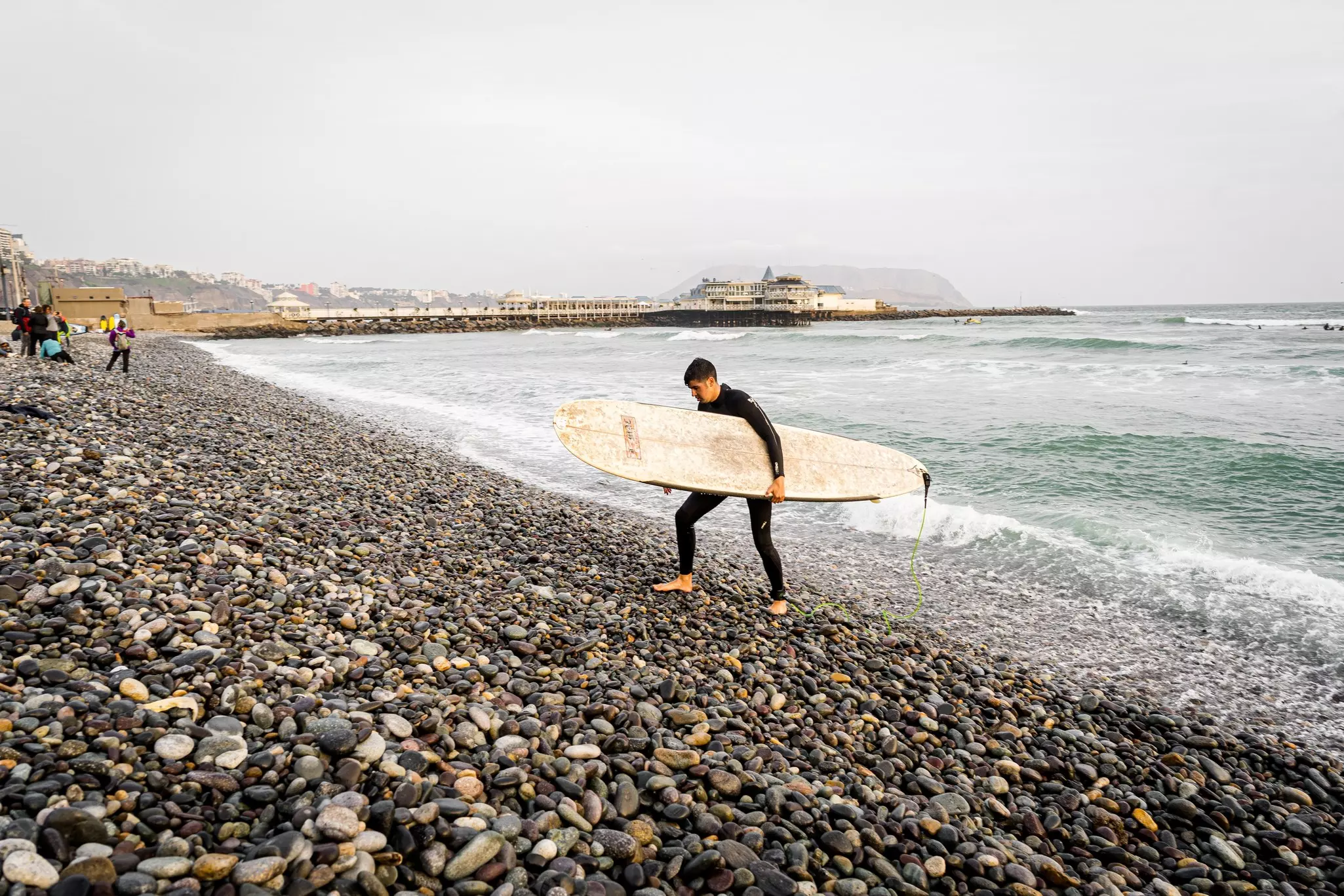 A surfer holding his board walks along a pebble beach in a city.