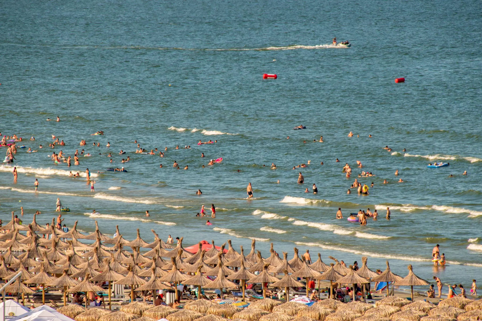 Sandy beach crowded with sunbathers under grass umbrellas with swimmers and waders in the distance.