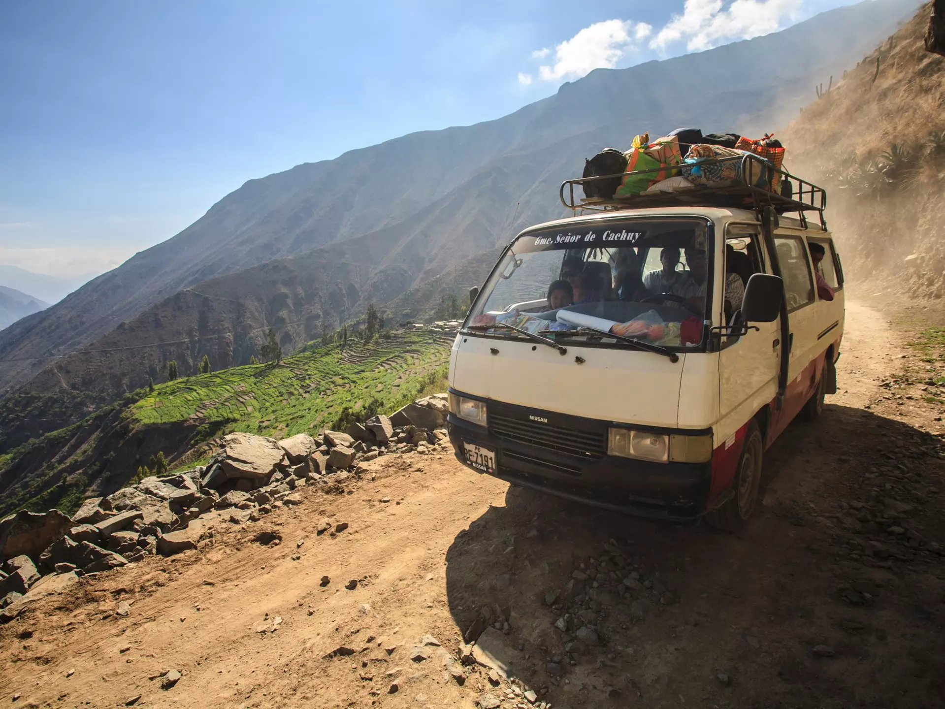 A small, old bus drives along a mountain dirt road in the Peruvian Andes mountain range. Next to the road is a steep drop leading down into a green valley.