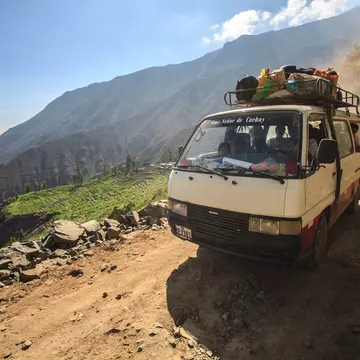 A small, old bus drives along a mountain dirt road in the Peruvian Andes mountain range. Next to the road is a steep drop leading down into a green valley.