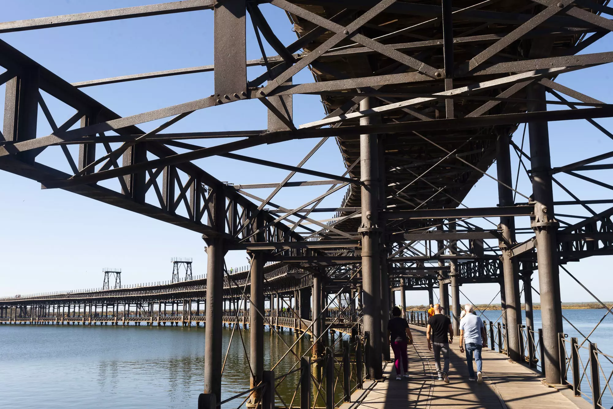 People walk along an iron pier structure that leads out over a waterway.