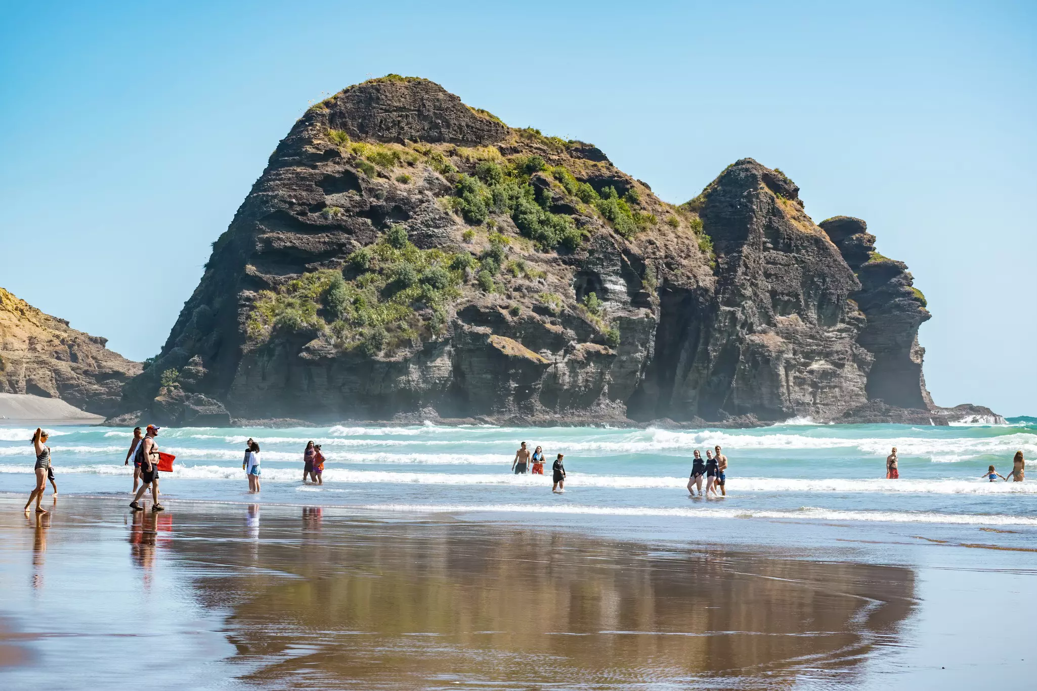 People swimming and walking at Piha beach, Auckland with Camel Rock cliff face ocean in background. Auckland  New Zealand - January 31 2020  License Type: media  Download Time: 2023-06-21T12:11:56.000Z  User:   Is Editorial: Yes  purchase_order: 56530  