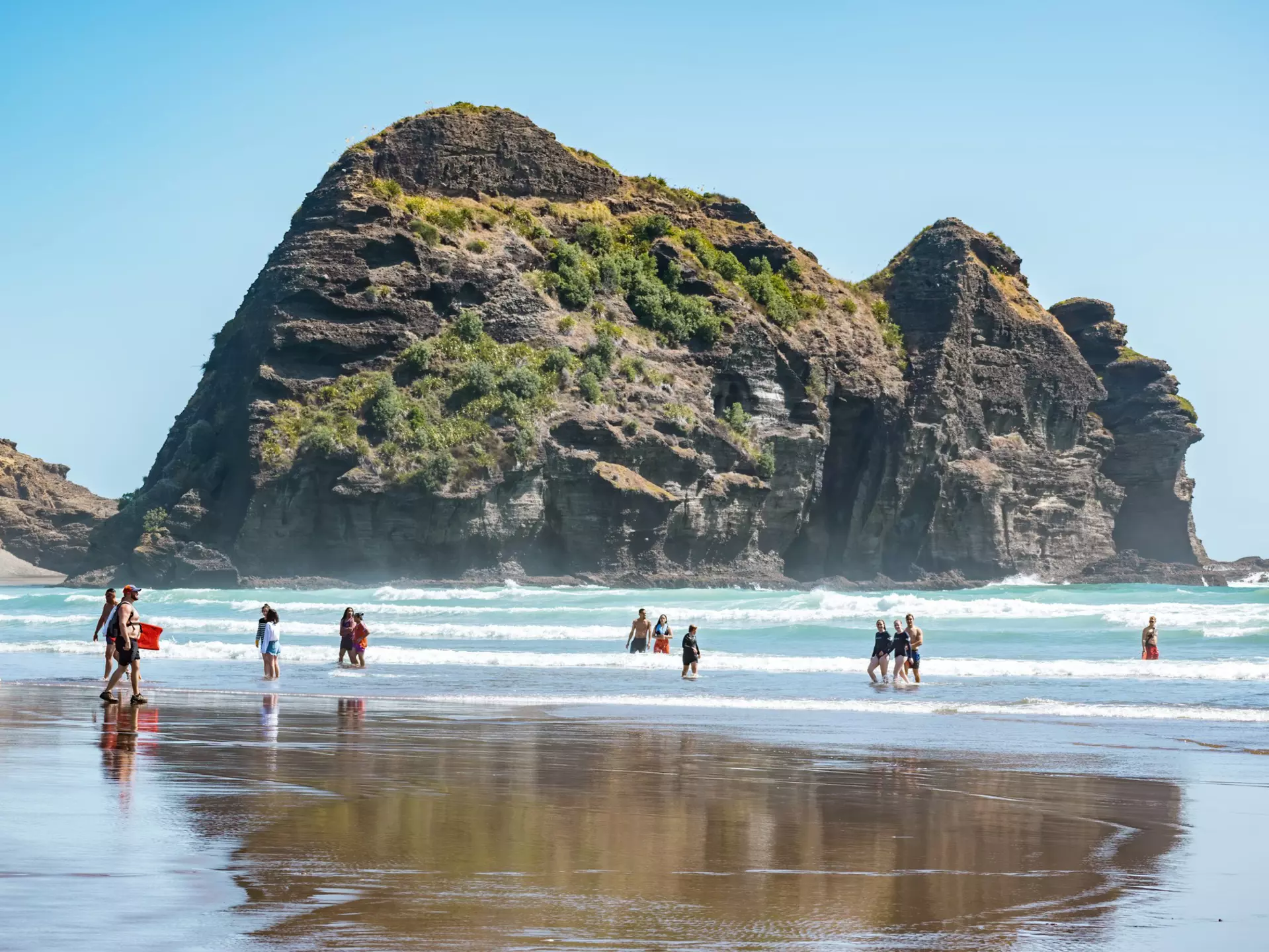 People paddling in the water at Piha beach with the Camel Rock in the background. 