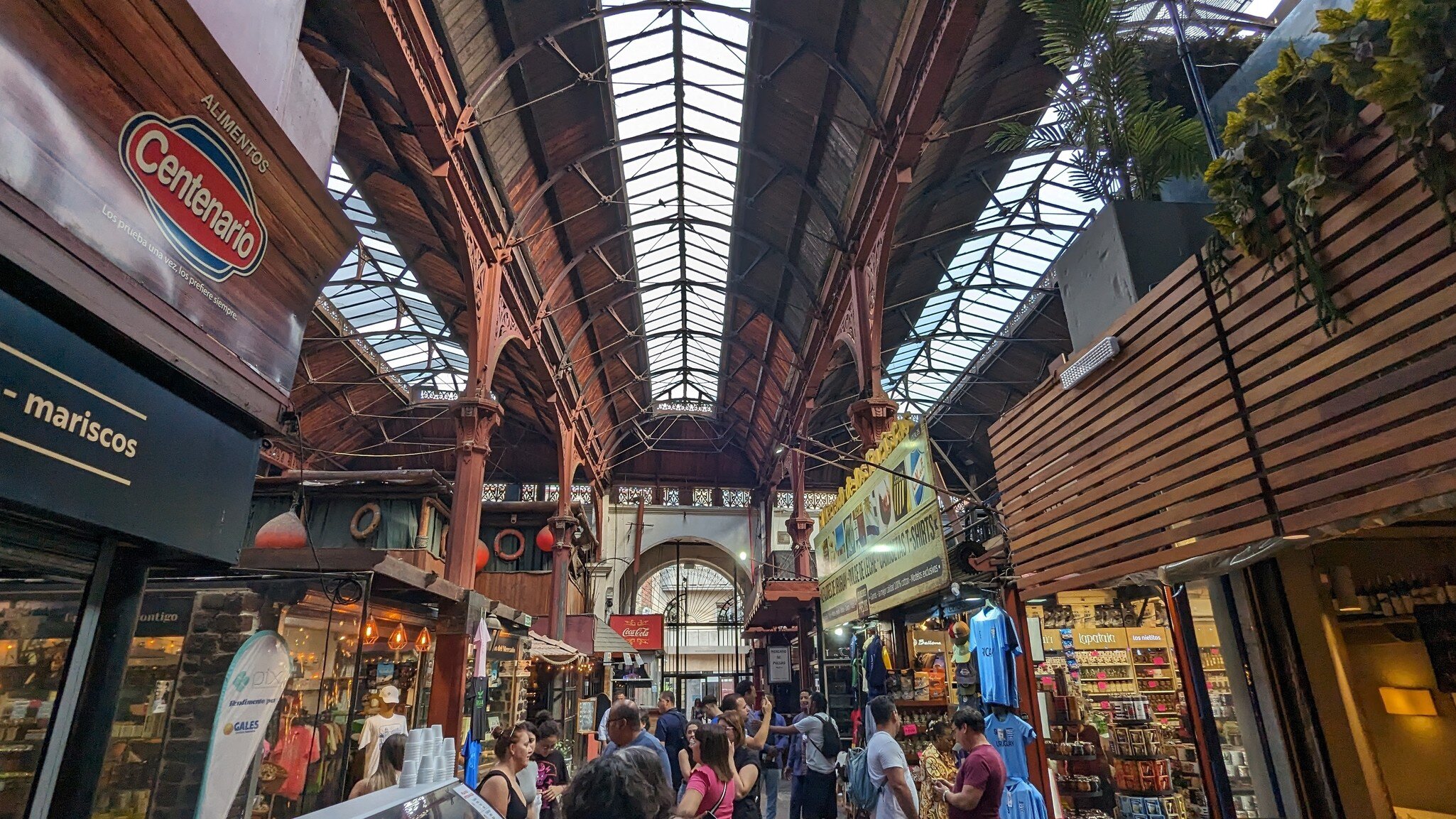 A busy market hall with a pitched wrought-iron and glass roof over timber-fronted stalls, a mariscos sign on the left and shoppers crowding the aisle.