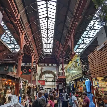 A busy market hall with a pitched wrought-iron and glass roof over timber-fronted stalls, a mariscos sign on the left and shoppers crowding the aisle.