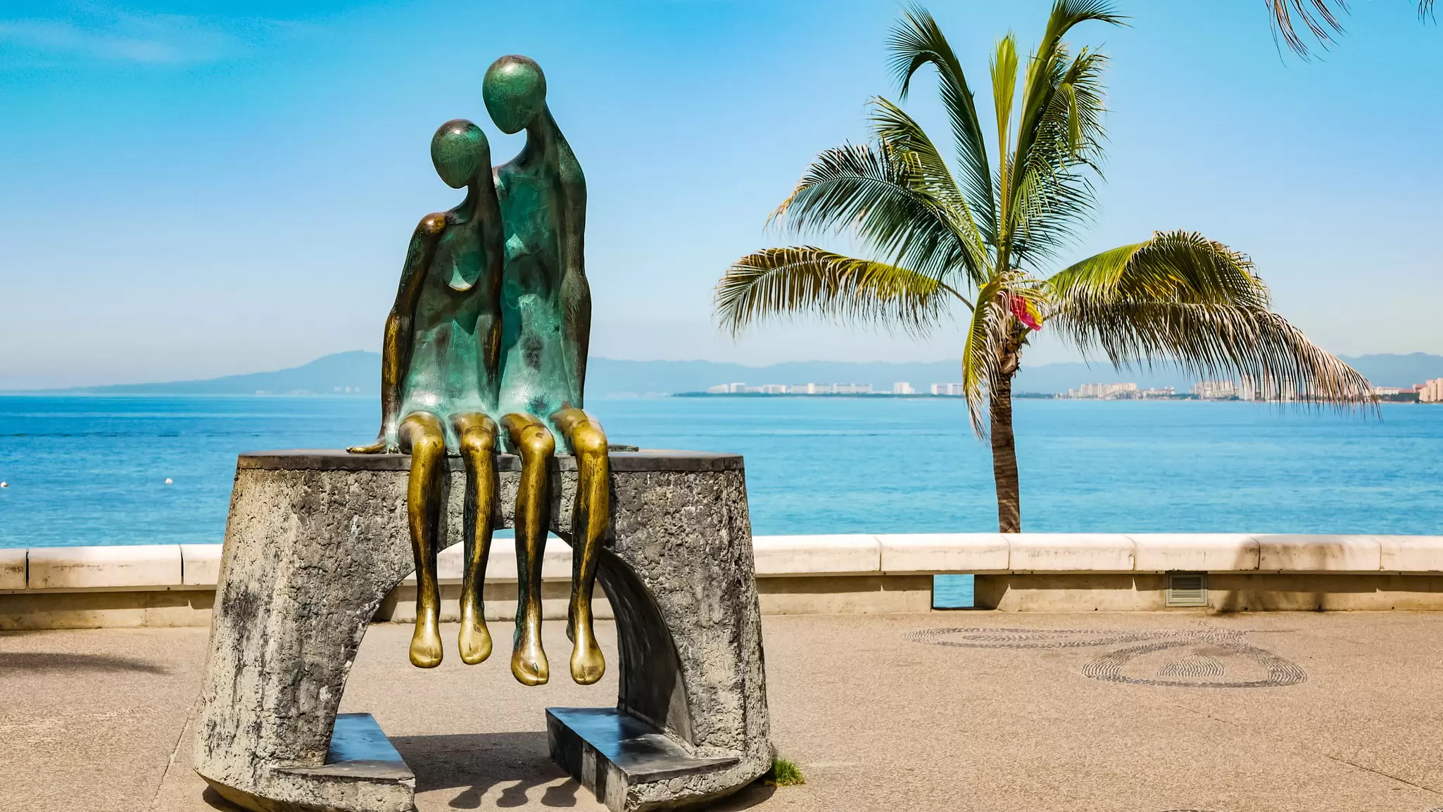 View of a bronze sculpture of a loving couple sitting side by side, with the ocean in the background, in Puerto Vallarta, Mexico.