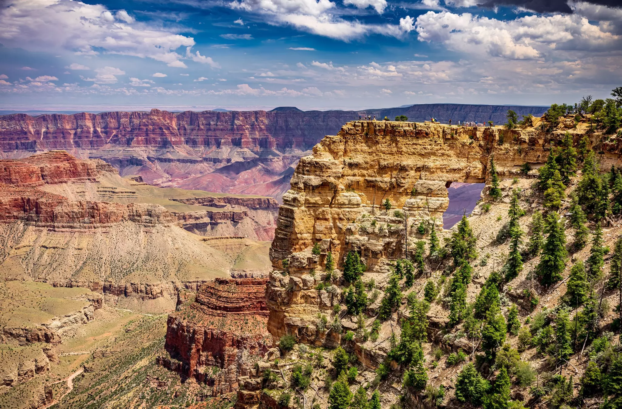 An opening in a rock formation at the edge of a large canyon with colorful striations in the rock.
