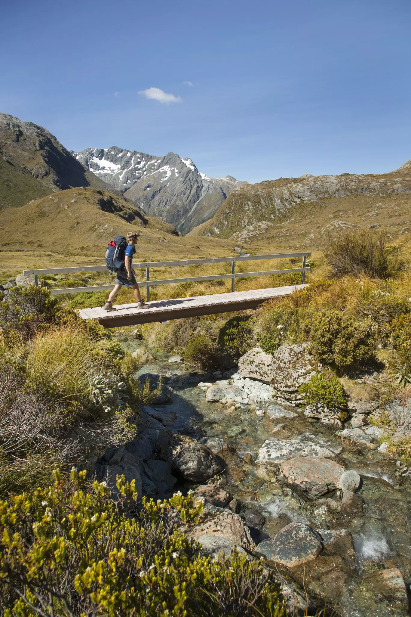 A hiker crosses a low wooden bridge over a creek in a mountainous landscape
