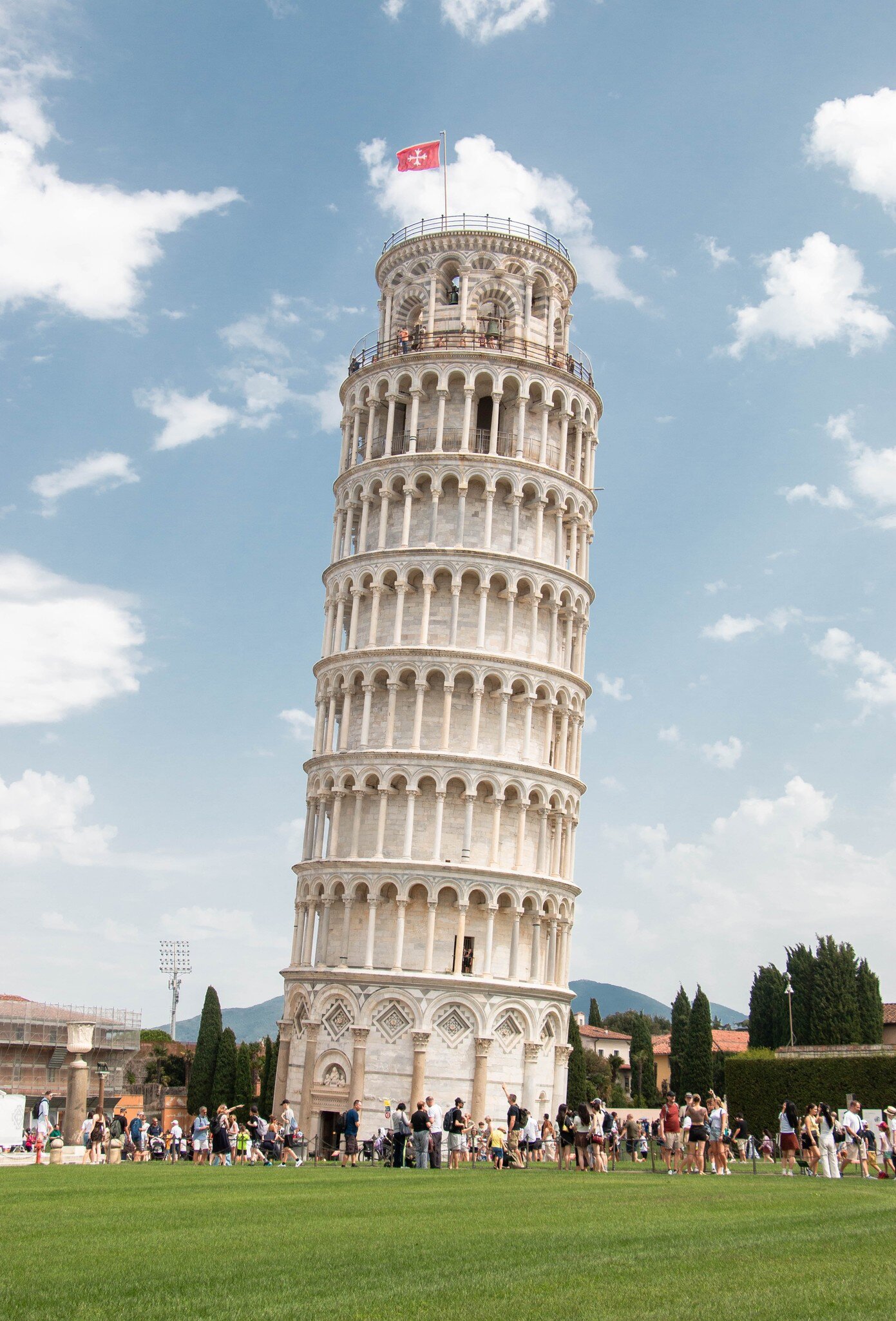 The Leaning Tower of Pisa with a red flag on top, tilting against a blue sky with white clouds, a crowd on the grass below.