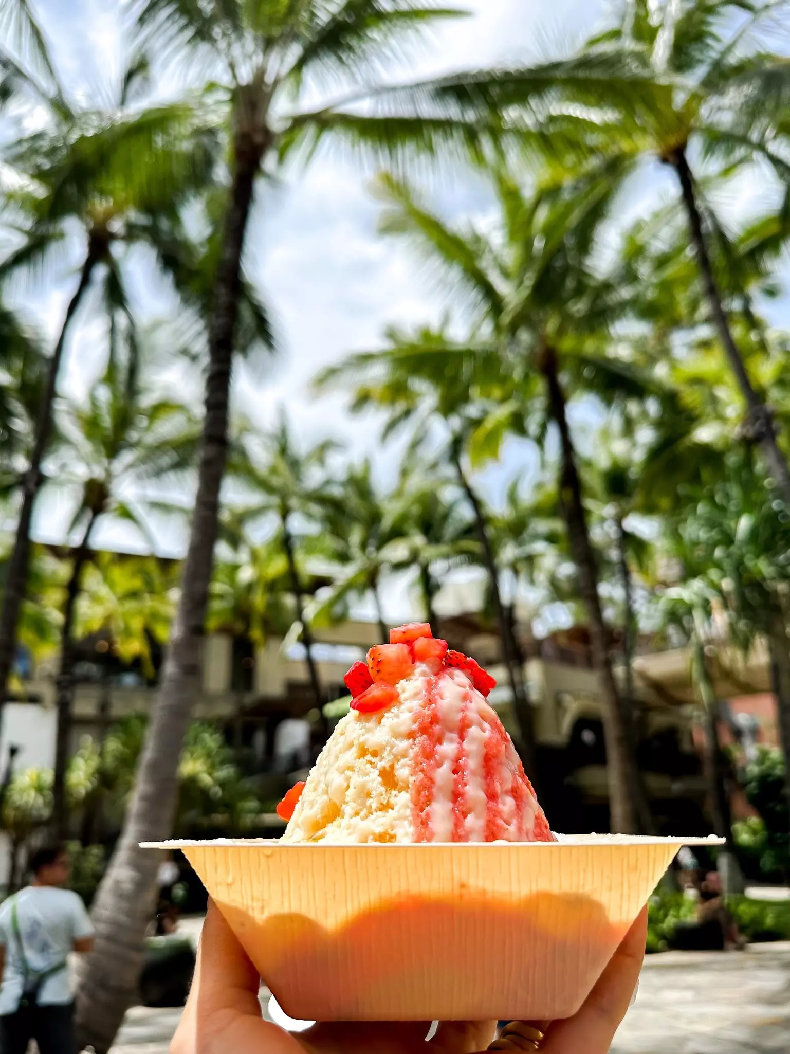 A cone filled with colorful shave ice held up under palm trees son Waikīkī Beach, Hawaiʻi
