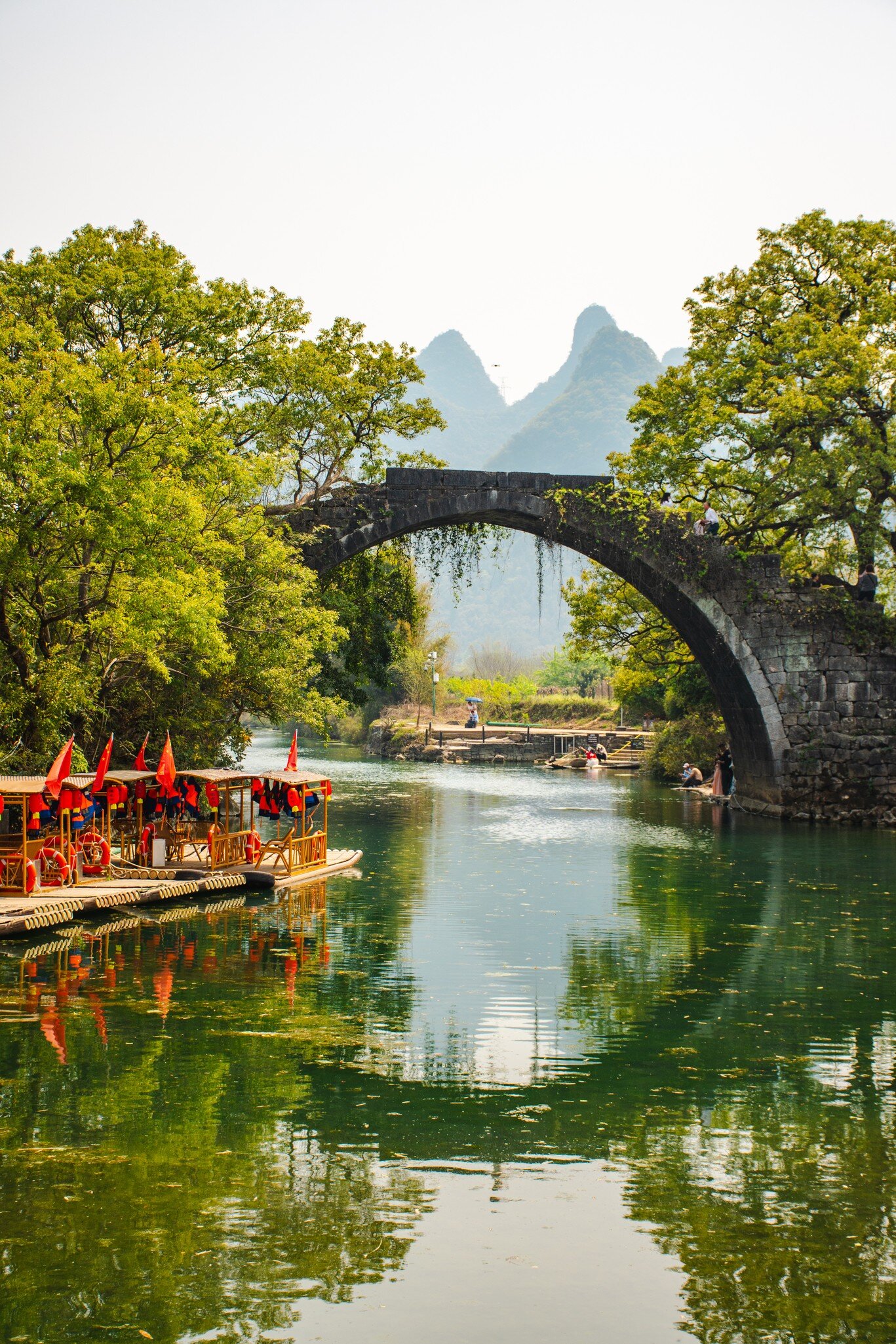 Bridge over by the river surrounded by green trees, in Yangshuo with mountain view in the background