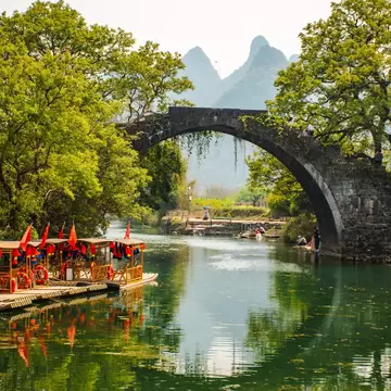 Bridge over by the river surrounded by green trees, in Yangshuo with mountain view in the background