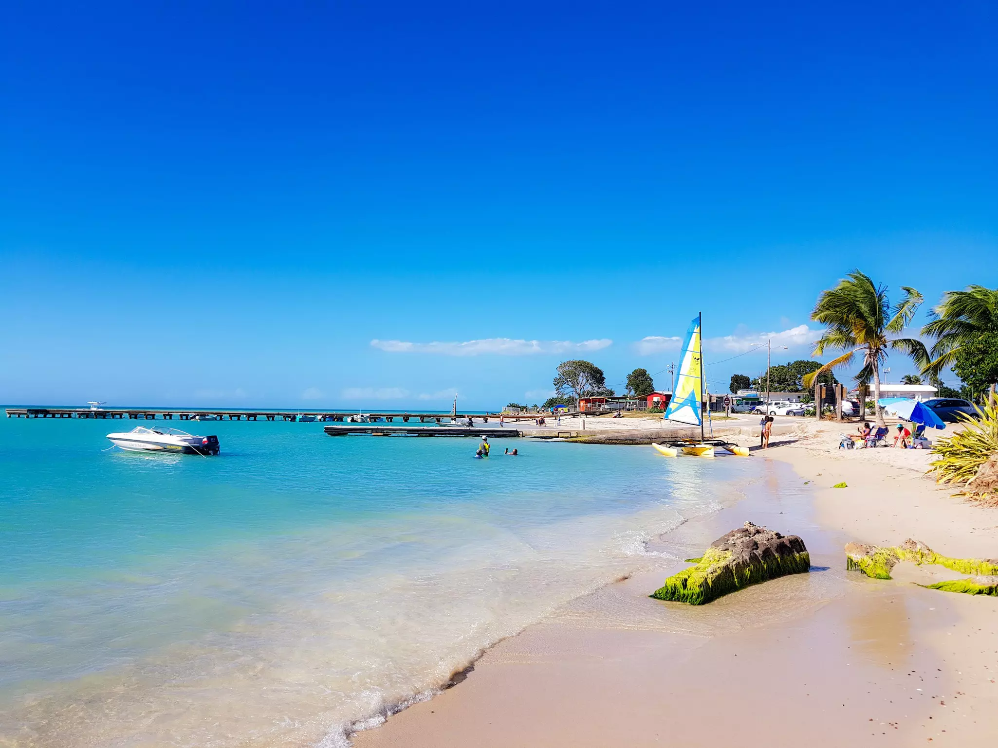 A sandy beach with turquoise ocean to the left and bright blue sky above. A catamaran and a few people are on the beach in the distance. A power boat is moored close off shore, and a long pier juts into the water in the far distance.