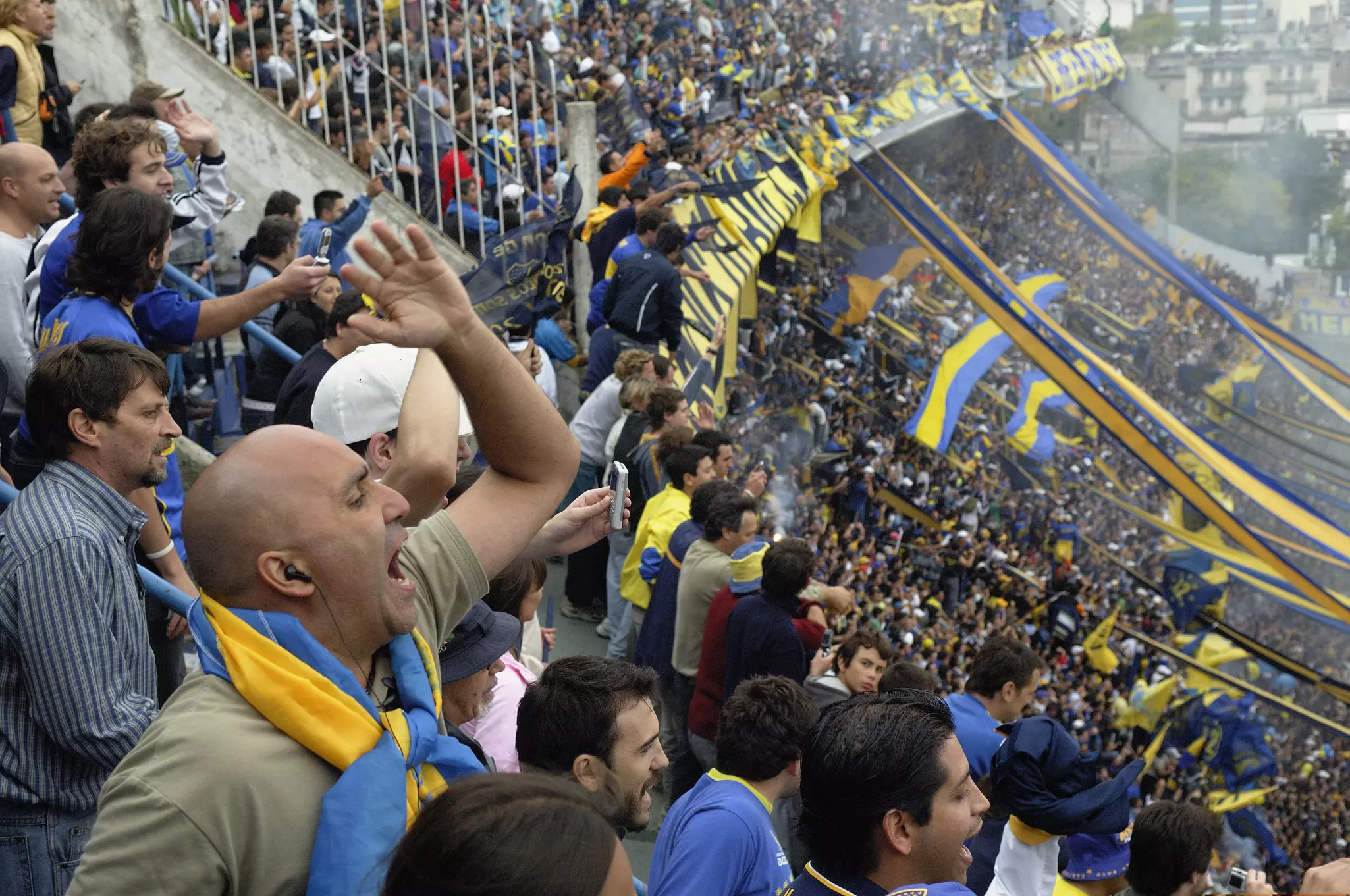 Spectators in home-team colors of blue and yellow cheer loudly and wave flags in the bleachers at a match in a packed stadium.