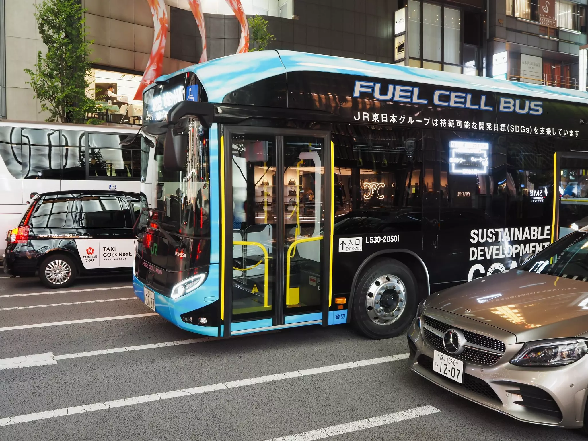 A blue and black hydrogen fuel cell bus and a black and white taxi going in opposite directions in traffic