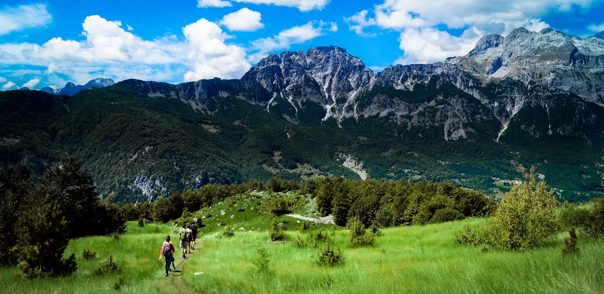 Hikers on the trail from Valbonë to Theth in Albania's Accursed Mountains, with rugged peaks framing the sky. 