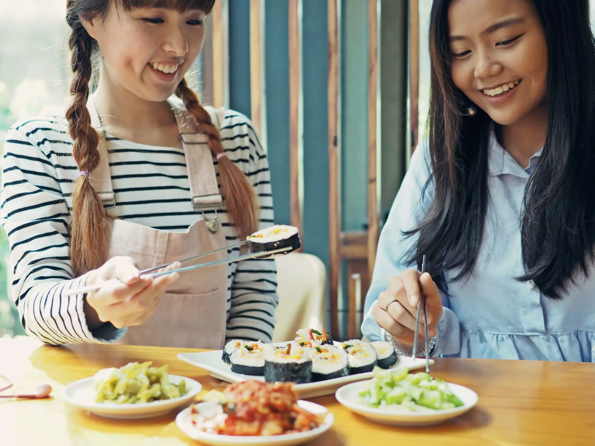 Asian women holding silver chopsticks to eat Korean seaweed rice roll with cheese or cheese kimbap and kimchi on wooden table.
