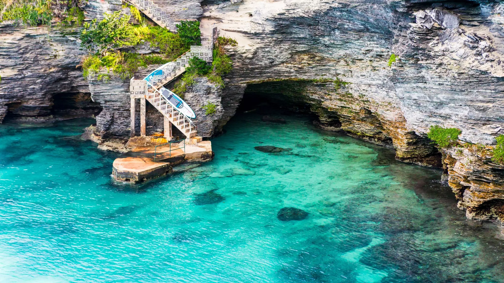 Wide shot of clear aqua ocean cove with rocks in the background and steps from the cliffs heading into the ocean. Two stand-up paddleboards are leaned against the stairway.
