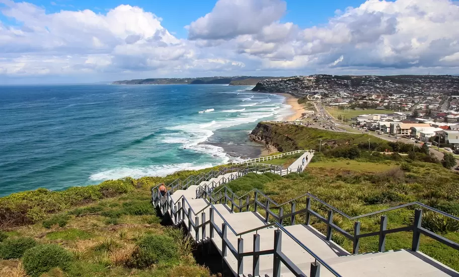 Steel stairway of Newcastle's Memorial Walk descending a grassy headland above surf, beaches and suburbs to the south