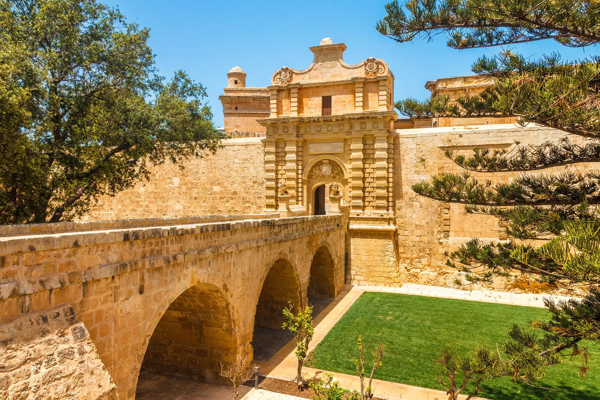 Entrance bridge and gate to Mdina, a fortified medieval city, with grass, trees and sky around