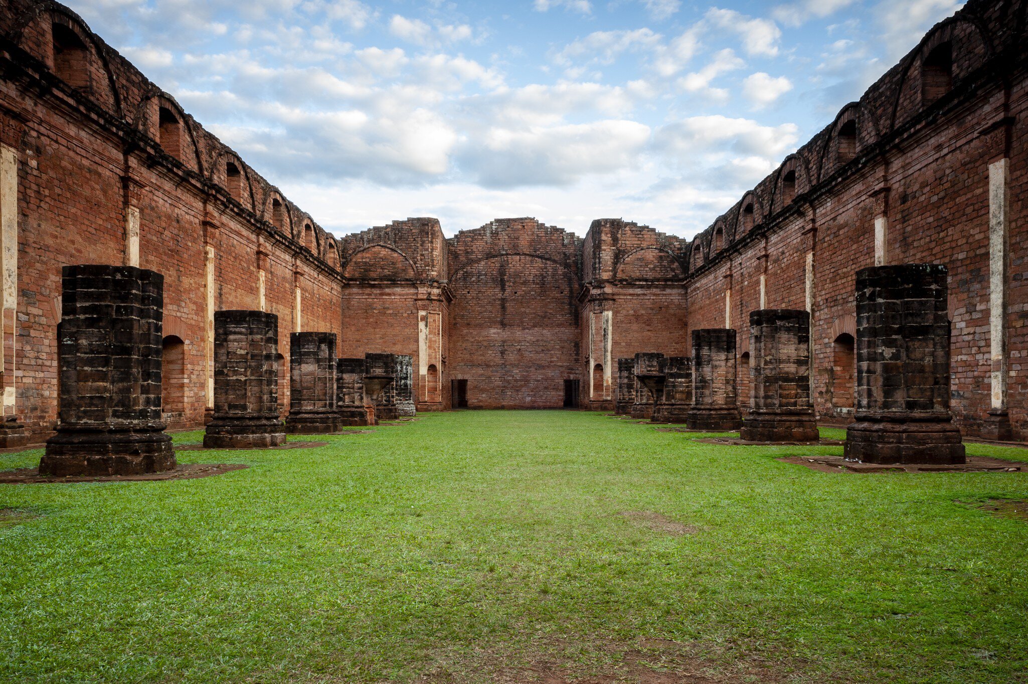 Grass floor between towering red walls, with squat stone column bases lining both sides under a cloudy blue sky