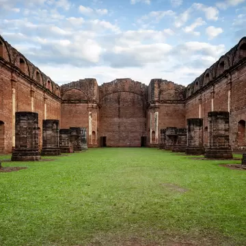 Grass floor between towering red walls, with squat stone column bases lining both sides under a cloudy blue sky